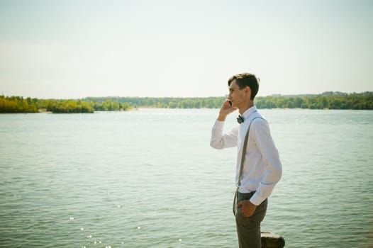 young skinny man, elegantly stylishly dressed in white shirt, gray trousers with suspenders and bow tie. Portrait of young guy talking on phone on background of river on warm sunny summer day