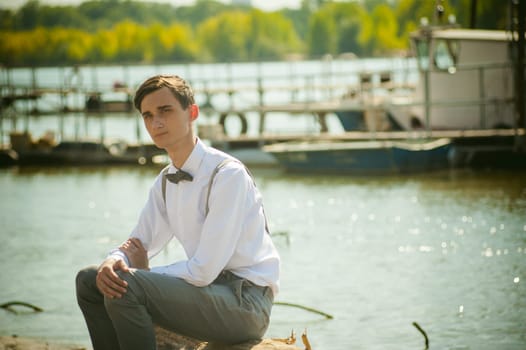 young skinny man, elegantly stylishly dressed in white shirt, gray trousers with suspenders and bow tie. Portrait young guy on pier on background of river and boats