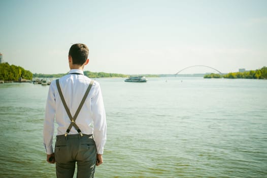 young skinny man, elegantly stylishly dressed in white shirt, gray trousers with suspenders and bow tie. Looking away, at the river water bridge and ship