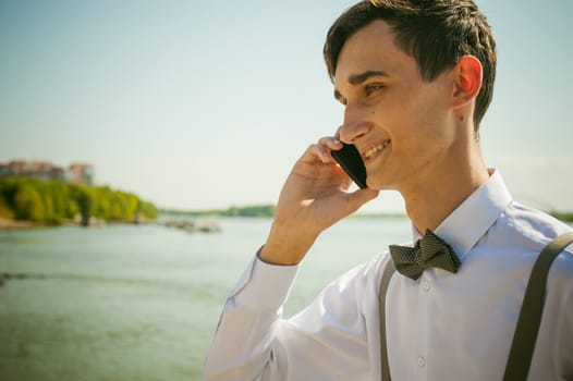 young skinny man, elegantly stylishly dressed in white shirt, gray trousers with suspenders and bow tie. Portrait of young guy talking on phone on background of river on warm sunny summer day