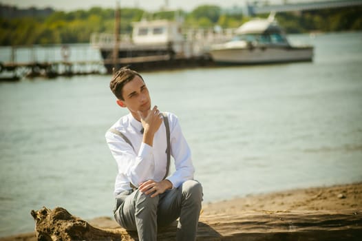 young skinny man, elegantly stylishly dressed in white shirt, gray trousers with suspenders and bow tie. Portrait young guy on pier on background of river and boats