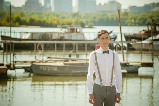 young skinny man, elegantly stylishly dressed in white shirt, gray trousers with suspenders and bow tie. Portrait young guy on pier on background of river and boats