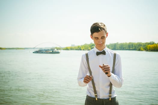 young skinny man, elegantly stylishly dressed in white shirt, gray trousers with suspenders and bow tie. Portrait of a guy on the background of a river with a passing boat at the bridge