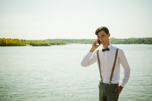 young skinny man, elegantly stylishly dressed in white shirt, gray trousers with suspenders and bow tie. Portrait of young guy talking on phone on background of river on warm sunny summer day