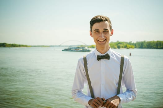 young skinny man, elegantly stylishly dressed in white shirt, gray trousers with suspenders and bow tie. Portrait of a guy on the background of a river with a passing boat at the bridge
