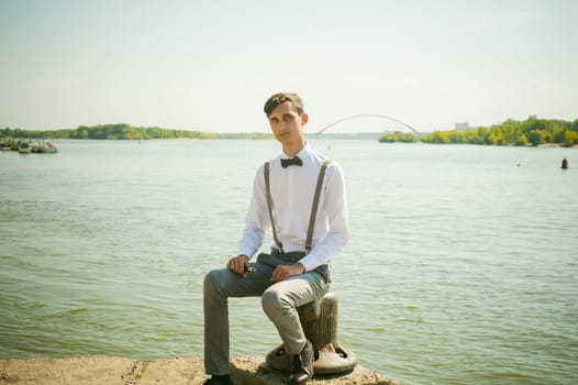 young skinny man, elegantly stylishly dressed in white shirt, gray trousers with suspenders and bow tie. guy sitting on a pier in the background of the river