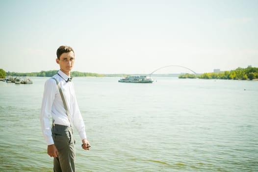 young skinny man, elegantly stylishly dressed in white shirt, gray trousers with suspenders and bow tie. Portrait of a guy on the background of a river with a passing boat at the bridge