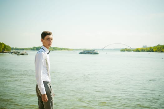 young skinny man, elegantly stylishly dressed in white shirt, gray trousers with suspenders and bow tie. Portrait of a guy on the background of a river with a passing boat at the bridge