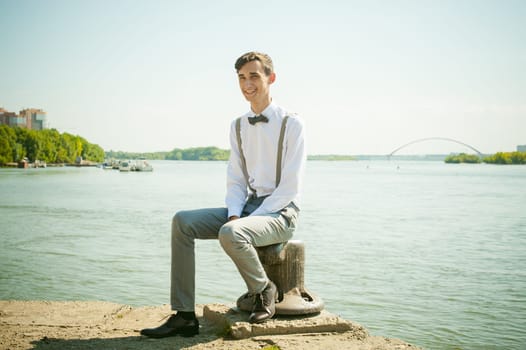 young skinny man, elegantly stylishly dressed in white shirt, gray trousers with suspenders and bow tie. guy sitting on a pier in the background of the river