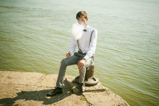 young skinny man, elegantly stylishly dressed in white shirt, gray trousers with suspenders and bow tie. guy smokes an electronic cigarette, smoke, sits on the pier, against background of river