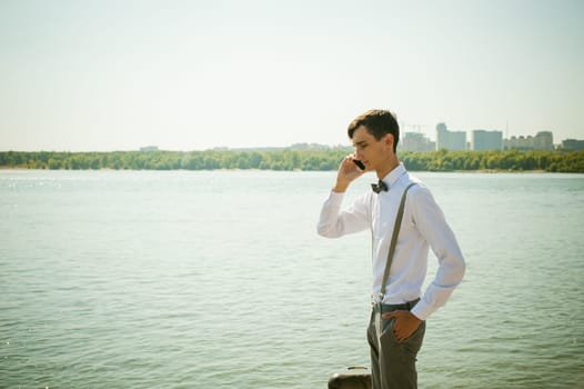 young skinny man, elegantly stylishly dressed in white shirt, gray trousers with suspenders and bow tie. Portrait of young guy talking on phone on background of river on warm sunny summer day