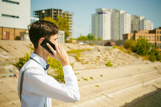 young skinny man, elegantly stylishly dressed in white shirt, gray trousers with suspenders and bow tie. Portrait of young guy talking on phone on background of river on warm sunny summer day