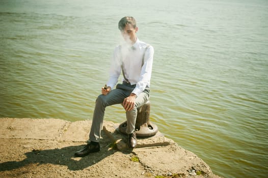 young skinny man, elegantly stylishly dressed in white shirt, gray trousers with suspenders and bow tie. guy smokes an electronic cigarette, smoke, sits on the pier, against background of river