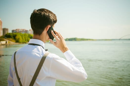 young skinny man, elegantly stylishly dressed in white shirt, gray trousers with suspenders and bow tie. Portrait of young guy talking on phone on background of river on warm sunny summer day