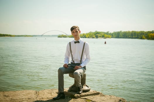 young skinny man, elegantly stylishly dressed in white shirt, gray trousers with suspenders and bow tie. guy sitting on a pier in the background of the river