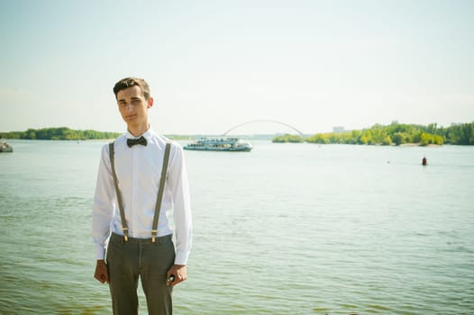 young skinny man, elegantly stylishly dressed in white shirt, gray trousers with suspenders and bow tie. Portrait of a guy on the background of a river with a passing boat at the bridge