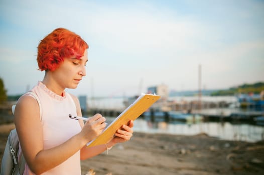 woman with dyed red hair in a pale pink dress with white backpack, signed important documents sitting outdoors near the river at sunset, against a boat mooring