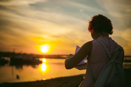 woman with dyed red hair in a pale pink dress with white backpack, signed important documents sitting outdoors near the river at sunset, against a boat mooring