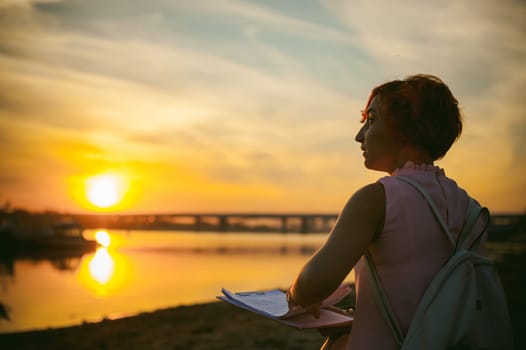 woman with dyed red hair in a pale pink dress with white backpack, signed important documents sitting outdoors near the river at sunset, against a boat mooring