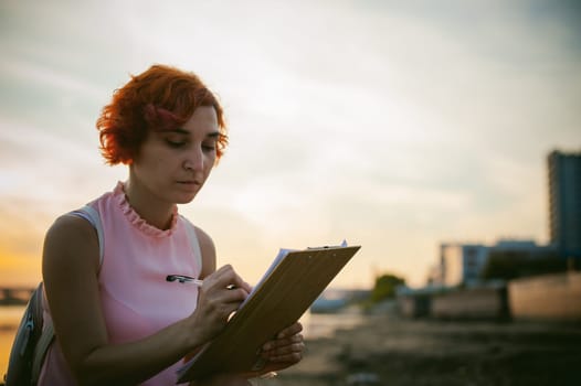 woman with dyed red hair in a pale pink dress with white backpack, signed important documents sitting outdoors near the river at sunset, against a boat mooring