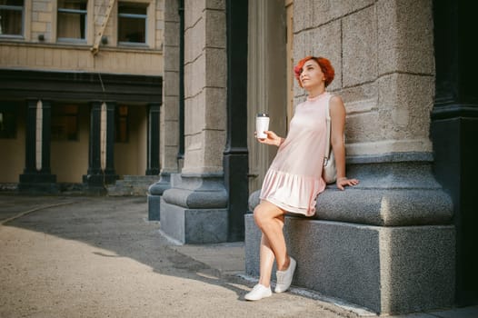 drinks coffee from a cup. woman with dyed red hair in a pale pink dress with a backpack white color drinking coffee, holding a paper cup of Take away, on a city street a warm summer day, behind, a huge exclusive doors