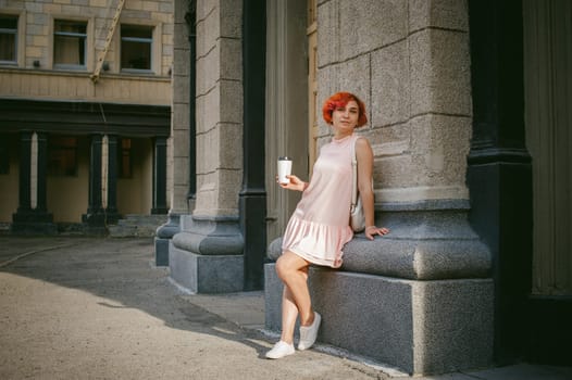 drinks coffee from a cup. woman with dyed red hair in a pale pink dress with a backpack white color drinking coffee, holding a paper cup of Take away, on a city street a warm summer day, behind, a huge exclusive doors