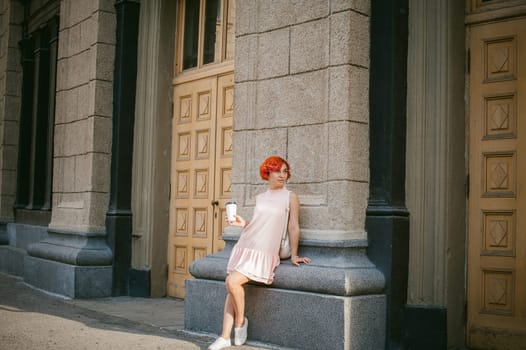 drinks coffee from a cup. woman with dyed red hair in a pale pink dress with a backpack white color drinking coffee, holding a paper cup of Take away, on a city street a warm summer day, behind, a huge exclusive doors
