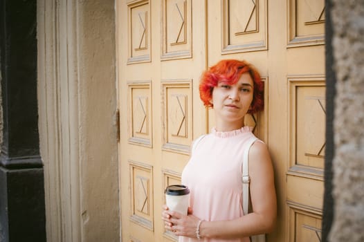 drinks coffee from a cup. woman with dyed red hair in a pale pink dress with a backpack white color drinking coffee, holding a paper cup of Take away, on a city street a warm summer day, behind, a huge exclusive doors