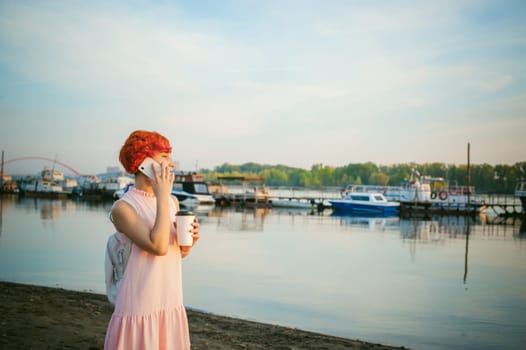 girl in pale pink dress with red hair and backpack walking along river bank, talking on the phone and drinking coffee from a cardboard cup, against backdrop of boats moored on a warm summer day