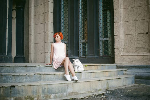 drinks coffee from a cup. woman with dyed red hair in a pale pink dress with a backpack white color drinking coffee, holding a paper cup of Take away, on a city street a warm summer day