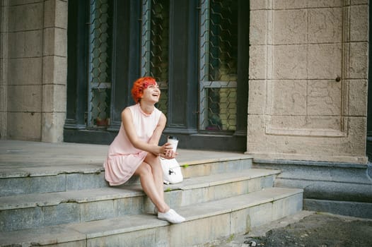 drinks coffee from a cup. woman with dyed red hair in a pale pink dress with a backpack white color drinking coffee, holding a paper cup of Take away, on a city street a warm summer day