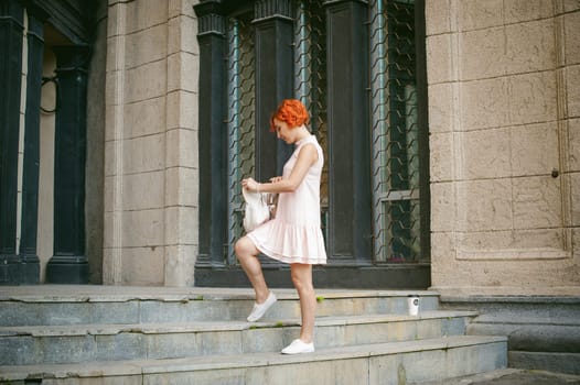 drinks coffee from a cup. woman with dyed red hair in a pale pink dress with a backpack white color drinking coffee, holding a paper cup of Take away, on a city street a warm summer day