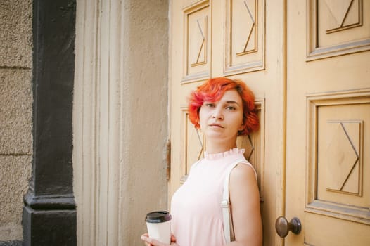 drinks coffee from a cup. woman with dyed red hair in a pale pink dress with a backpack white color drinking coffee, holding a paper cup of Take away, on a city street a warm summer day, behind, a huge exclusive doors