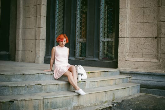 drinks coffee from a cup. woman with dyed red hair in a pale pink dress with a backpack white color drinking coffee, holding a paper cup of Take away, on a city street a warm summer day