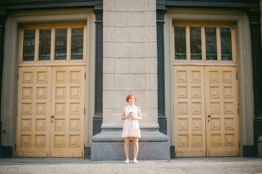 drinks coffee from a cup. woman with dyed red hair in a pale pink dress with a backpack white color drinking coffee, holding a paper cup of Take away, on a city street a warm summer day, behind, a huge exclusive doors