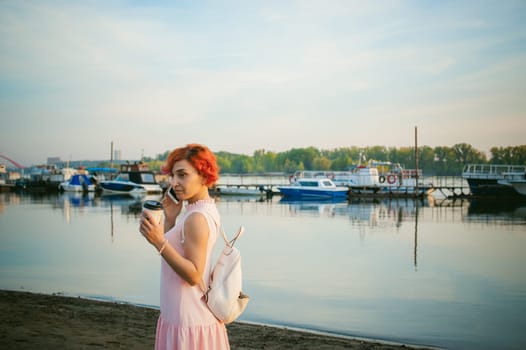girl in pale pink dress with red hair and backpack walking along river bank, talking on the phone and drinking coffee from a cardboard cup, against backdrop of boats moored on a warm summer day