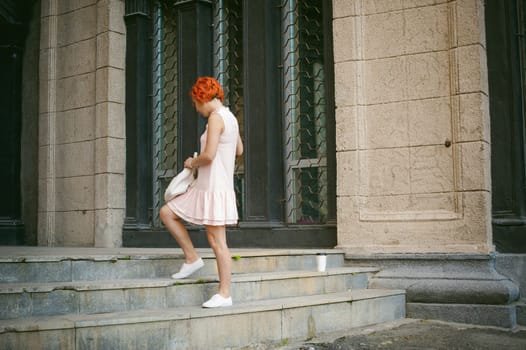 drinks coffee from a cup. woman with dyed red hair in a pale pink dress with a backpack white color drinking coffee, holding a paper cup of Take away, on a city street a warm summer day