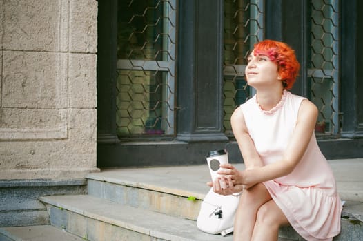 drinks coffee from a cup. woman with dyed red hair in a pale pink dress drinking coffee, holding a paper cup of coffee to go, sitting on the steps outside a summer day.