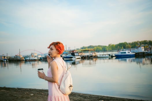 girl in pale pink dress with red hair and backpack walking along river bank, talking on the phone and drinking coffee from a cardboard cup, against backdrop of boats moored on a warm summer day