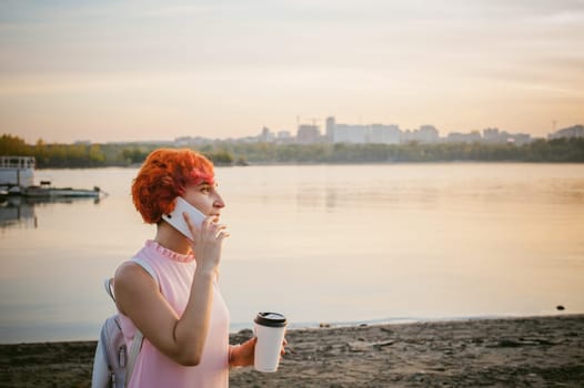 girl in pale pink dress with red hair and backpack walking along river bank, talking on the phone and drinking coffee from a cardboard cup, against backdrop of boats moored on a warm summer day