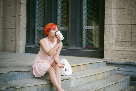 drinks coffee from a cup. woman with dyed red hair in a pale pink dress drinking coffee, holding a paper cup of coffee to go, sitting on the steps outside a summer day.
