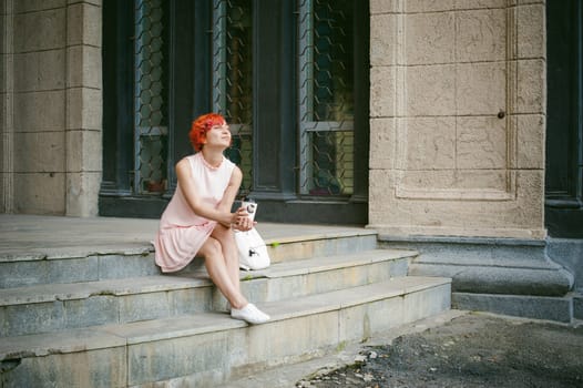 drinks coffee from a cup. woman with dyed red hair in a pale pink dress with a backpack white color drinking coffee, holding a paper cup of Take away, on a city street a warm summer day