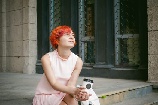 drinks coffee from a cup. woman with dyed red hair in a pale pink dress drinking coffee, holding a paper cup of coffee to go, sitting on the steps outside a summer day.