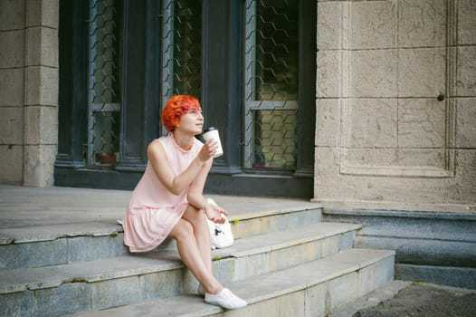 drinks coffee from a cup. woman with dyed red hair in a pale pink dress drinking coffee, holding a paper cup of coffee to go, sitting on the steps outside a summer day.