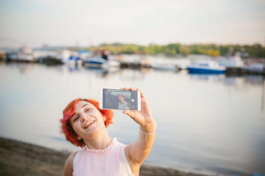 girl in pale pink dress with red hair and backpack walking along river bank, pictures of themselves on their mobile camera phone, against backdrop of boats moored on a warm summer day