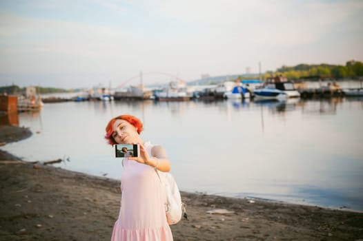 girl in pale pink dress with red hair and backpack walking along river bank, pictures of themselves on their mobile camera phone, against backdrop of boats moored on a warm summer day