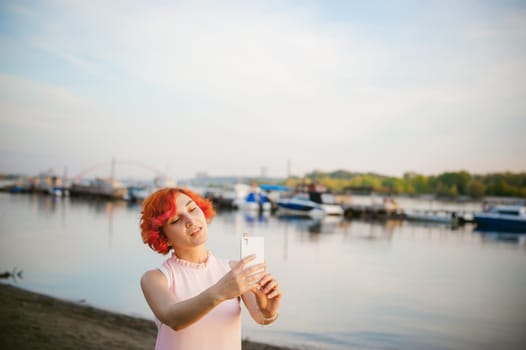 girl in pale pink dress with red hair and backpack walking along river bank, pictures of themselves on their mobile camera phone, against backdrop of boats moored on a warm summer day