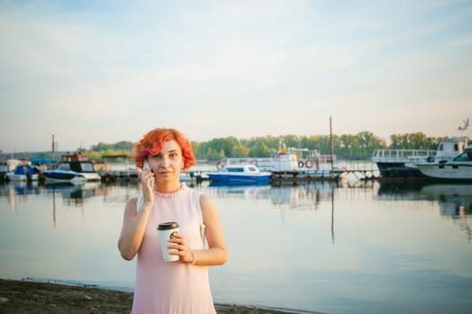 girl in pale pink dress with red hair and backpack walking along river bank, talking on the phone and drinking coffee from a cardboard cup, against backdrop of boats moored on a warm summer day