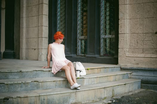 drinks coffee from a cup. woman with dyed red hair in a pale pink dress with a backpack white color drinking coffee, holding a paper cup of Take away, on a city street a warm summer day