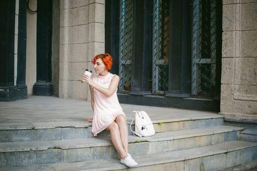 drinks coffee from a cup. woman with dyed red hair in a pale pink dress drinking coffee, holding a paper cup of coffee to go, sitting on the steps outside a summer day.